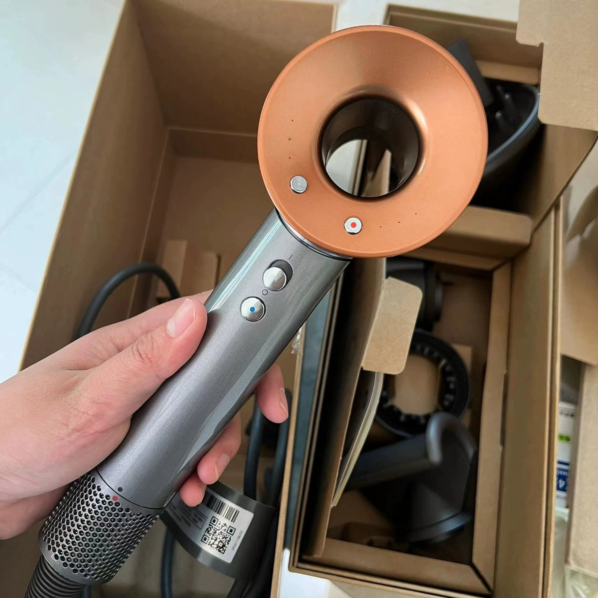Hand holding a cordless hair dryer with copper-colored circular attachment and gray handle with control buttons, displayed