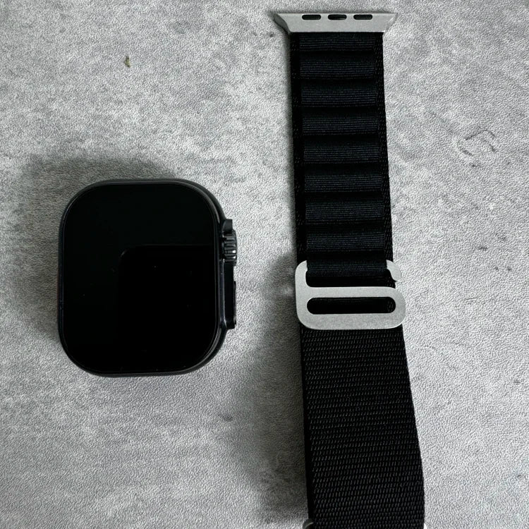 Black smartwatch with square face and dark screen next to black fabric watch band with white connector clasp on gray