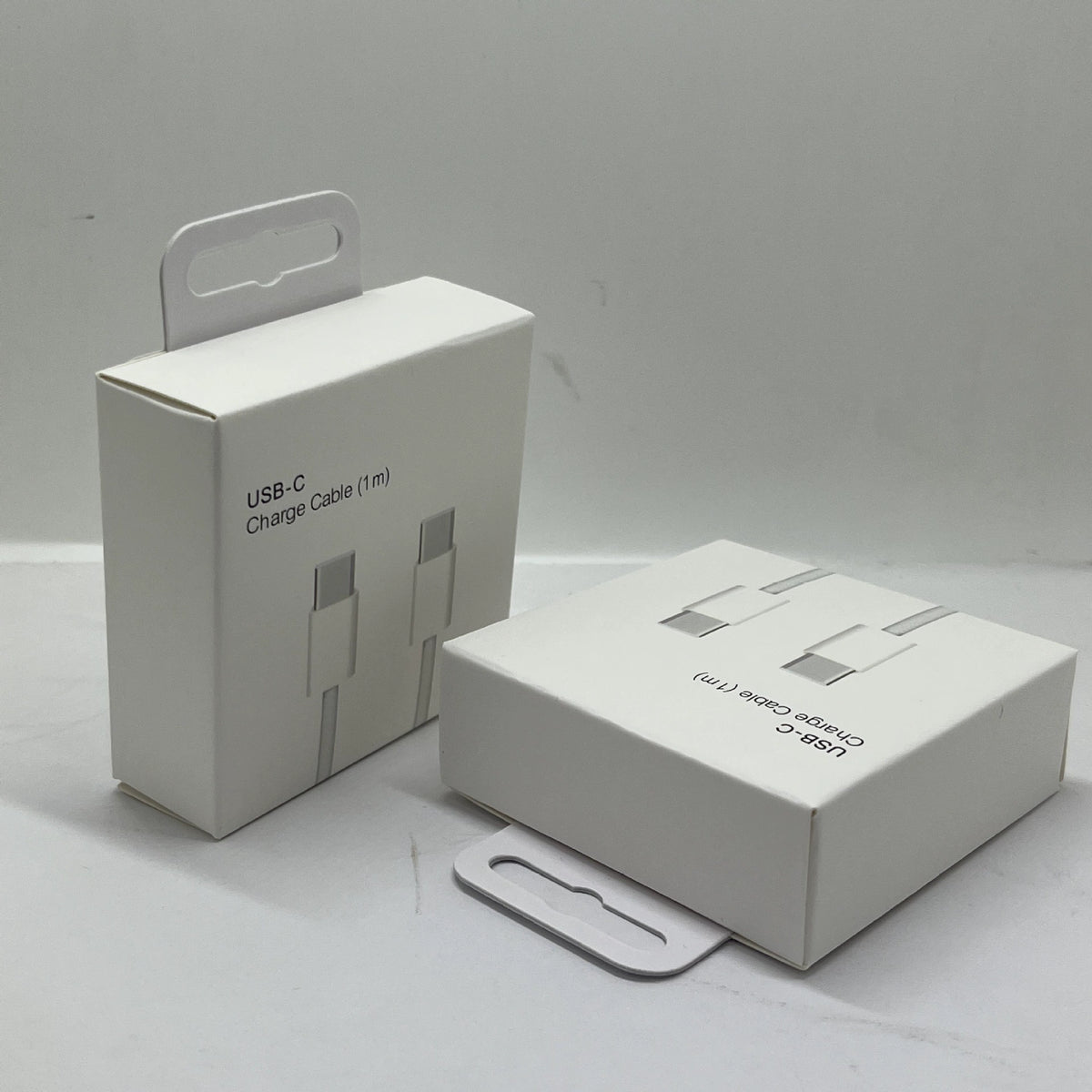Two white cardboard boxes with handles displayed on a gray surface; the left box is labeled "USB-C Charge Cable (1m)" with