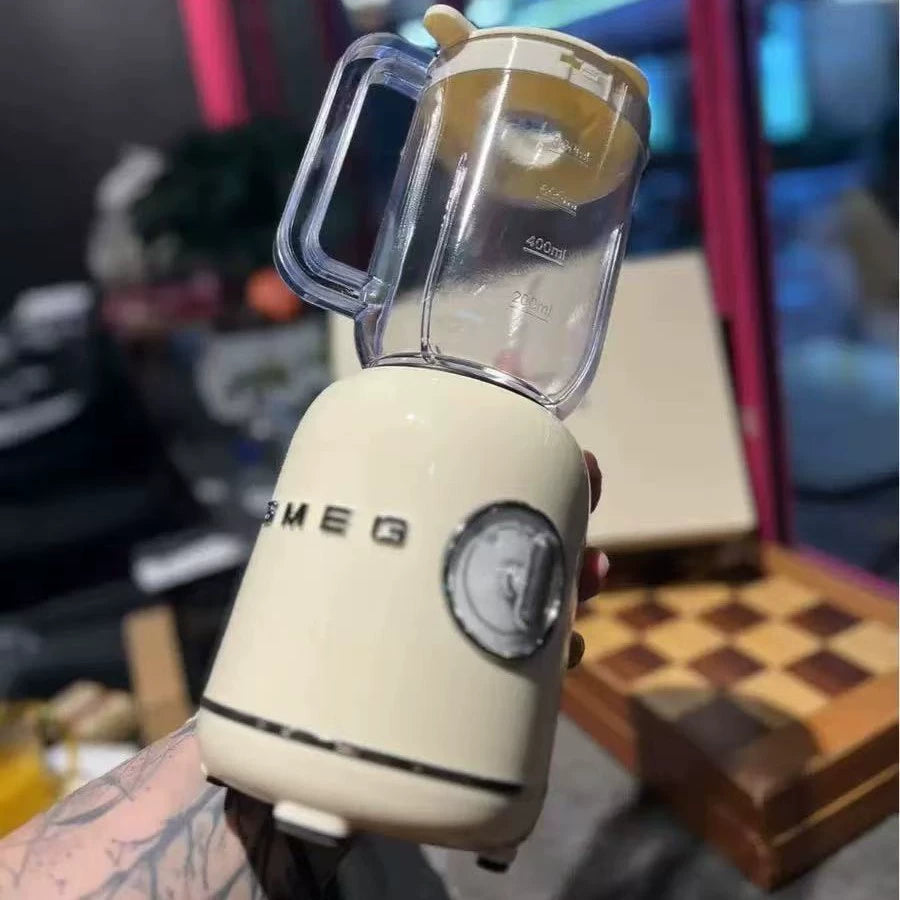 Cream-colored SMEG blender with clear glass pitcher on kitchen counter, held by hand against blurred colorful background.